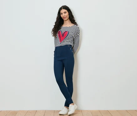 Frau mit dunklen, lockigen Haaren, gekleidet in ein gestreiftes Langarmshirt mit einem roten Herz darauf, marineblaue Hose und weiße Turnschuhe, posiert vor einer weißen Wand.