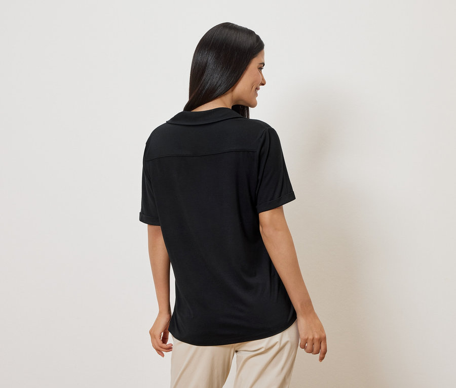 Frau von hinten in schwarzem Poloshirt und beigen Hosen vor weißem Hintergrund.