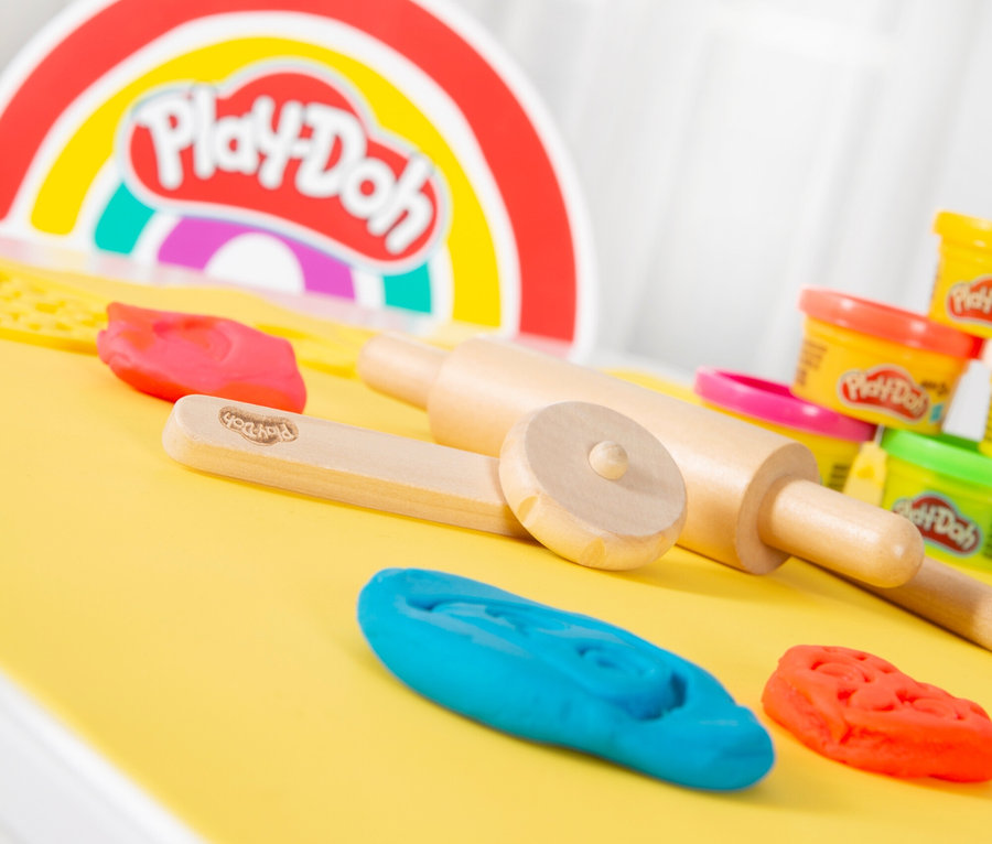 Play-Doh-Knete, Holzroller und Förmchen liegen auf einer gelben Matte vor einem Play-Doh-Regenbogen.