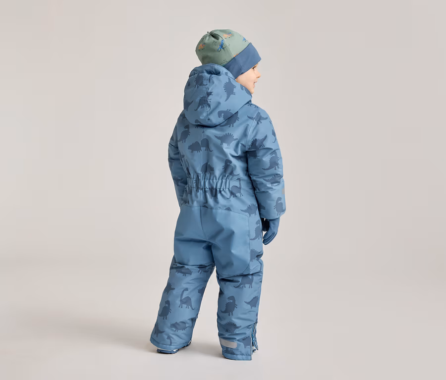 Ein Kind mit dem Rücken zur Kamera trägt einen blauen Schneeanzug mit Dinosaurier-Muster und eine grüne Mütze mit Dinosauriern.