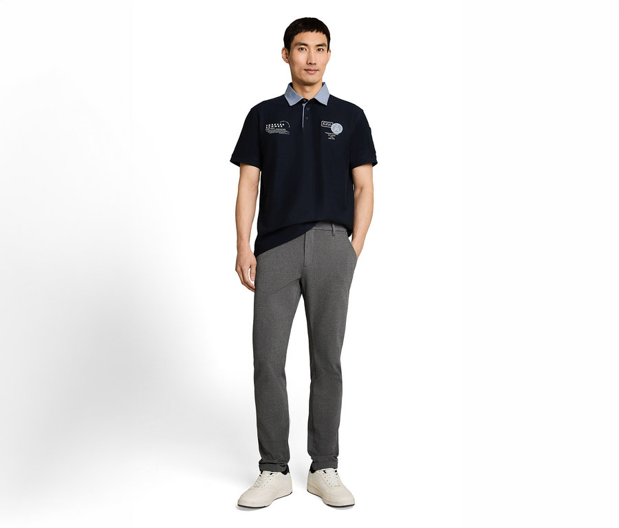 Ein Mann steht frontal, trägt ein dunkelblaues Poloshirt mit hellblauem Kragen und weißer Stickerei, eine graue Hose und helle Sneaker. Seine Hände stecken in den Hosentaschen.