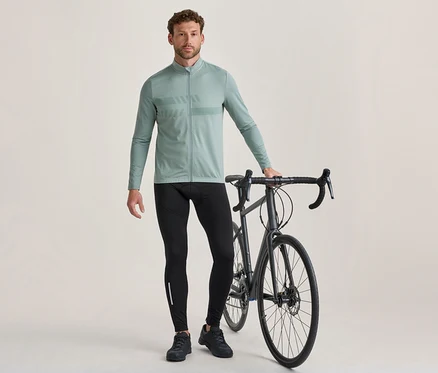 Ein Mann steht neben einem dunkelgrauen Rennrad und hält dessen Lenker. Er trägt ein hellgrünes, langärmeliges Radtrikot mit Brustmuster, schwarze Radhosen mit einem reflektierenden Streifen und schwarze Radschuhe.