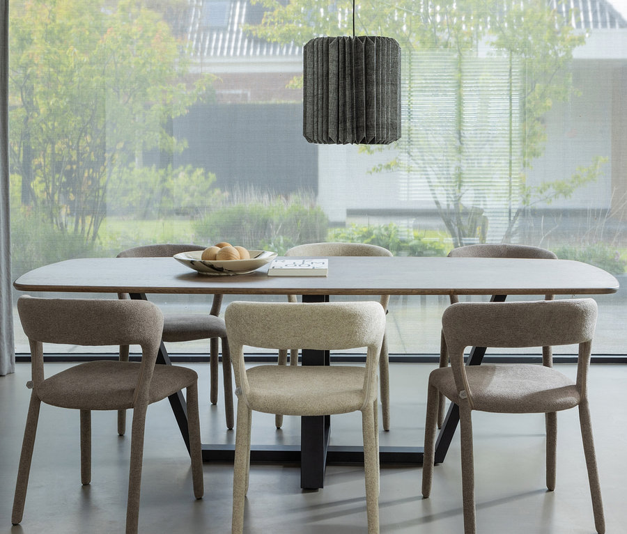 Ein modernes Esszimmer mit einem dunklen, rechteckigen Tisch, umgeben von sechs gepolsterten Stühlen in Hell- und Dunkelbeige. Eine graue, plissierte Pendelleuchte hängt über dem Tisch, auf dem eine dekorative Schale mit runden Objekten steht. Ein großes Fenster mit transparenten Rollos blickt auf einen Garten.
