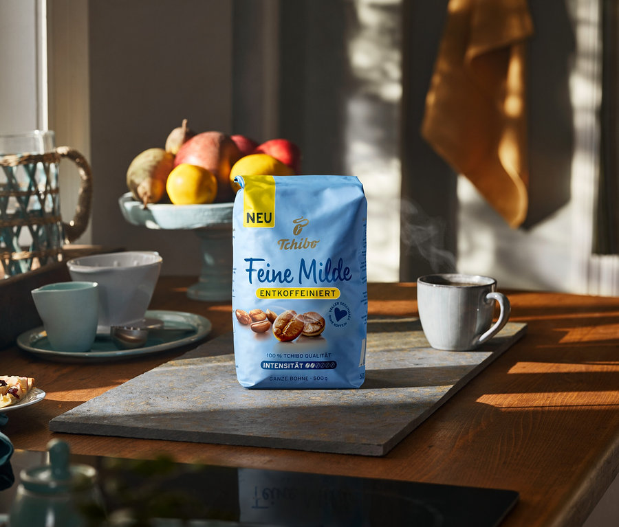 Eine blaue Packung Tchibo Feine Milde entkoffeinierten Kaffees steht auf einer grauen Matte auf einer Holzküchenarbeitsplatte, neben einer dampfenden Tasse Kaffee. Im Hintergrund sind eine Obstschale mit Granatäpfeln und Zitronen sowie ein Teller mit Gebäck zu sehen.