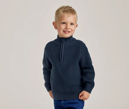 Lächelnder kleiner Junge mit blonden Haaren in einem marineblauen Rippstrickpullover mit halbem Reißverschluss und blauen Jeans.