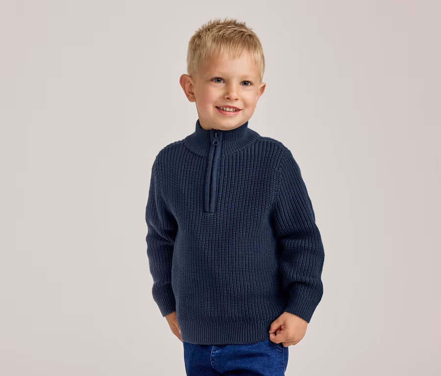 Lächelnder kleiner Junge mit blonden Haaren in einem marineblauen Rippstrickpullover mit halbem Reißverschluss und blauen Jeans.