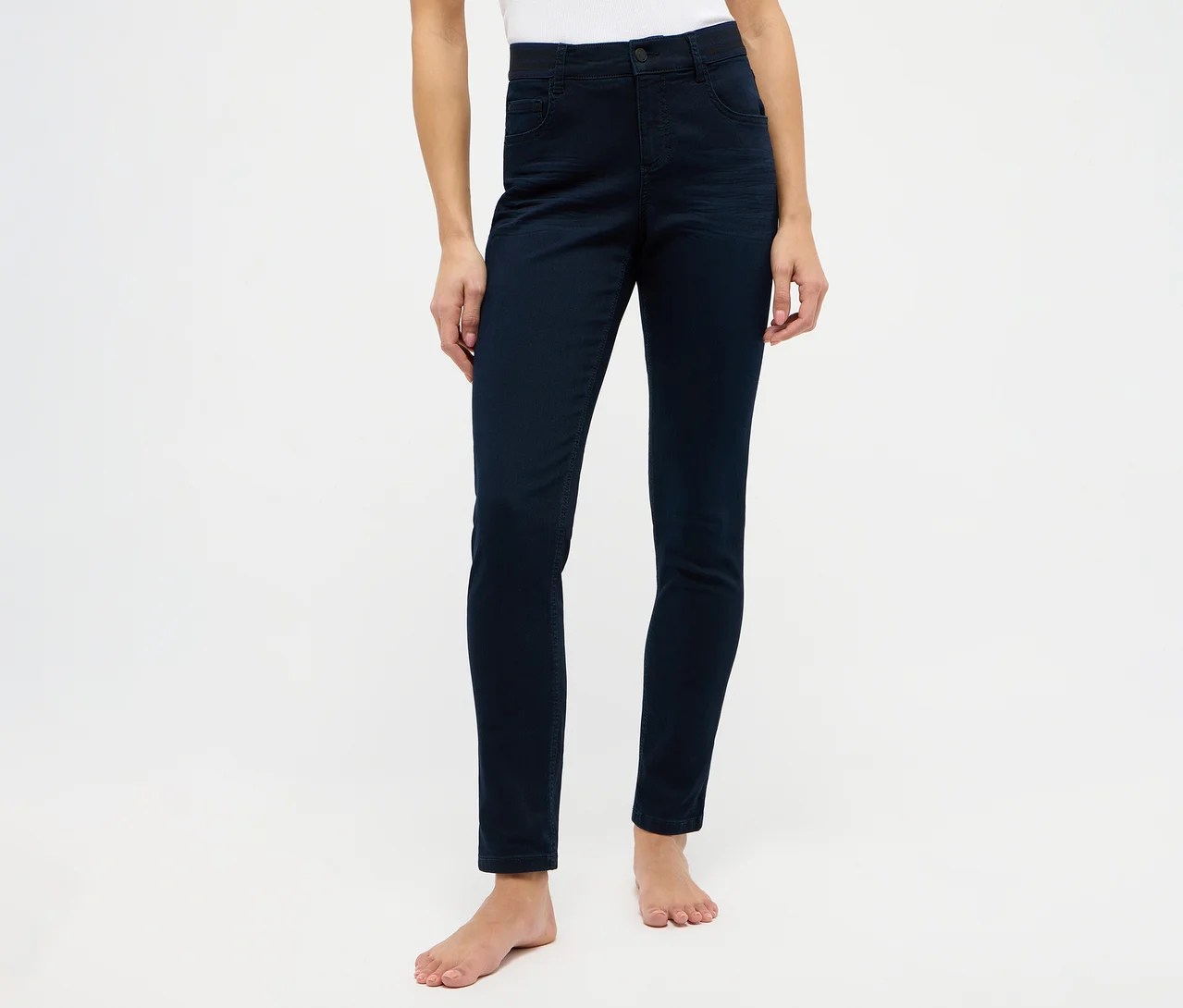 Person trägt dunkelblaue Skinny Jeans und ein weißes Oberteil, barfuß vor weißem Hintergrund stehend.
