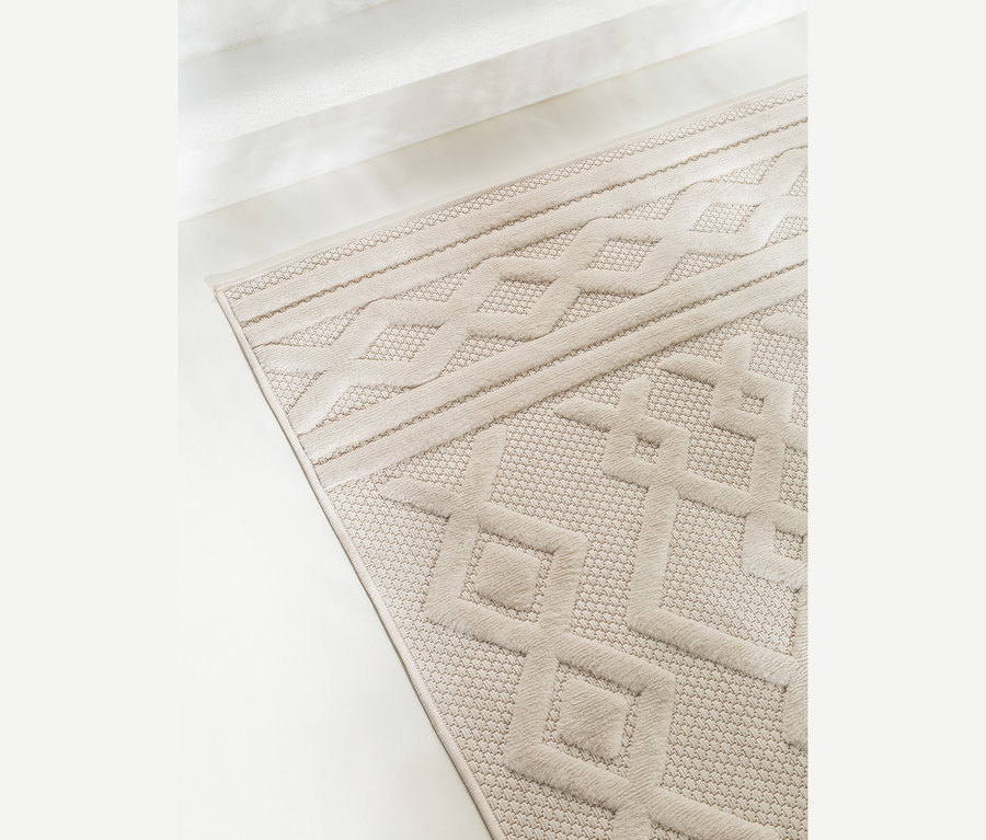 Heller beiger Teppich mit erhabenen geometrischen Mustern, darunter Zopf- und Rautenformen, auf hellem Boden neben einer weißen Wand.