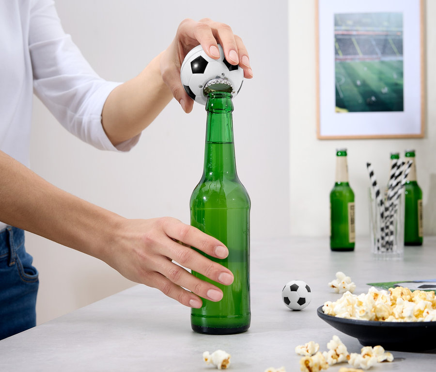 Eine Person öffnet eine grüne Bierflasche mit einem Fußball-Flaschenöffner. Auf dem Tisch befinden sich Popcorn und andere Flaschen.