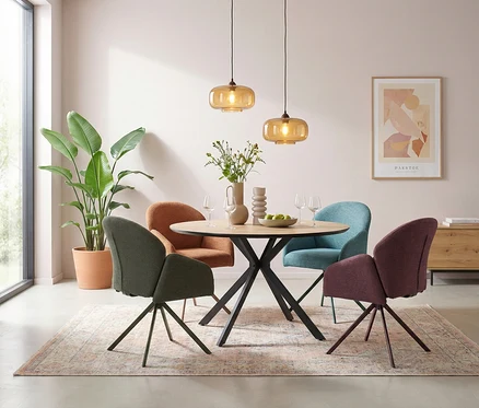 Ein modern eingerichtetes Esszimmer mit einem runden Holztisch und vier bunten Stühlen in Grün, Orange, Hellblau und Lila. Über dem Tisch hängen zwei bernsteinfarbene Pendelleuchten, und auf dem Tisch stehen eine Vase mit Blumen, eine Schale mit Äpfeln und Weingläser. Ein großer Topf mit einer Pflanze steht neben einem Fenster, und ein gemusterter Teppich liegt unter dem Esstisch.