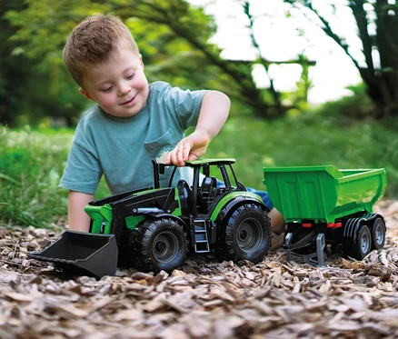 Ein lächelnder Junge spielt draußen auf Holzspänen mit einem grünen WORXX Traktor Deutz-Fahr Agrotron mit Frontlader und Anhänger.