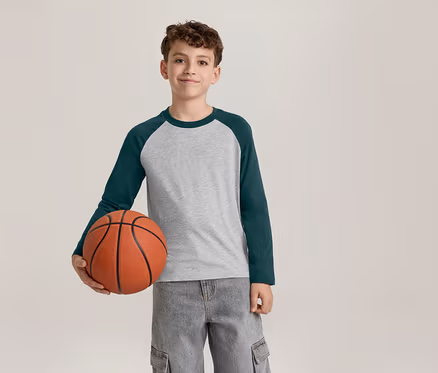 Ein lächelnder Junge in einem grau-dunkelgrünen Langarmshirt und grauen Cargohosen hält einen Basketball in seiner linken Hand.