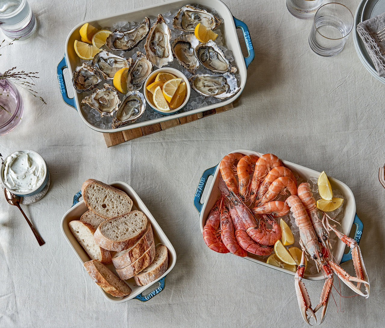 Drei weiße STAUB Auflaufformen mit blauen Griffen auf einem Tisch. Eine Form enthält Austern auf Eis mit Zitronenspalten, eine andere Garnelen und Langusten auf Eis mit Zitrone, und die dritte geschnittenes Brot.