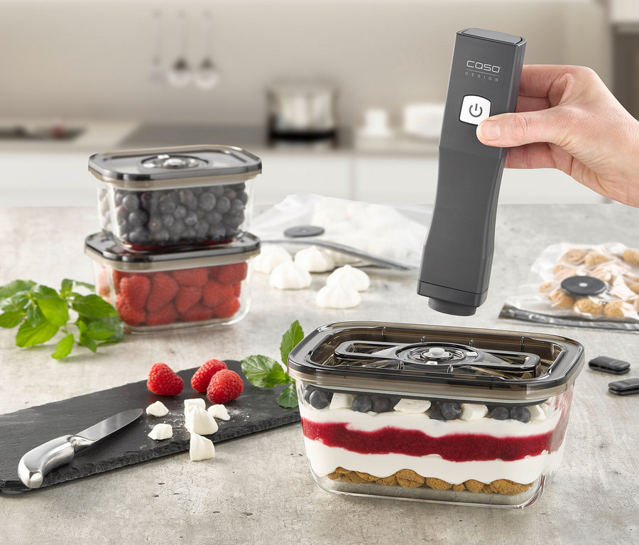 Eine Hand hält einen CASO Vacu OneTouch Handvakuumierer über einem Behälter mit Schichtdessert. Daneben stehen Behälter mit Beeren und eine Schieferplatte mit Himbeeren.