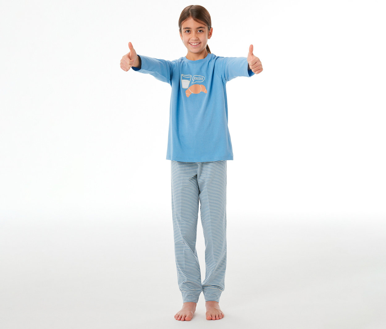 Lächelndes Mädchen in einem blauen SCHIESSER Kinder-Pyjama-Oberteil mit Croissant- und Milch-Aufdruck und blau-weiß gestreiften Pyjama-Hosen, das beide Daumen hochhält.