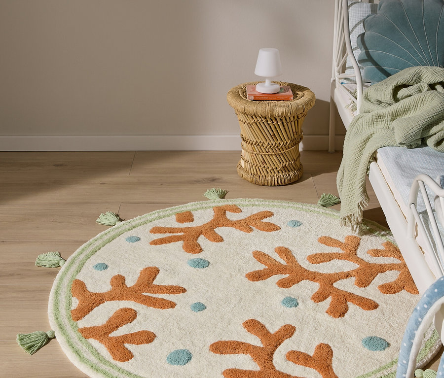 Kinderzimmer mit einem runden, cremefarbenen Teppich, der orange Korallenformen und blaue Punkte zeigt, einem grünen Rand mit Quasten, einem weißen Metallbett und einem Beistelltisch aus Korbgeflecht mit einer Lampe.