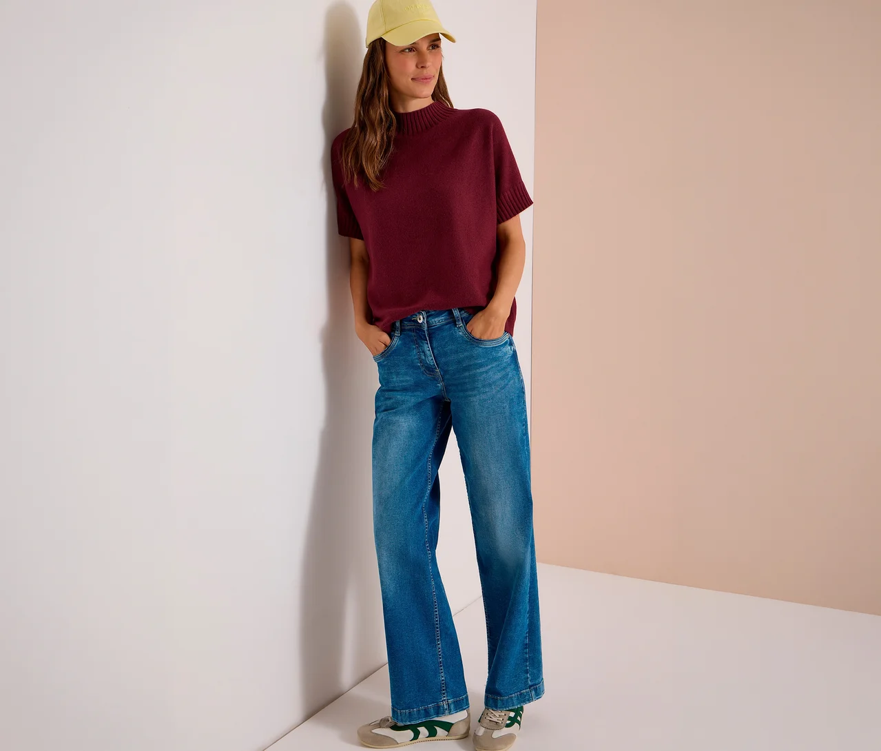 Eine Frau trägt einen bordeauxroten CECIL Strickpullover, weite Jeans und eine gelbe Baseballkappe, während sie mit den Händen in den Taschen an einer Wand lehnt.