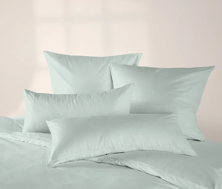 Salbeigrüne Jersey-Kopfkissenbezüge und ein passender Bettbezug liegen ordentlich auf einem Bett vor einer hellrosa Wand.