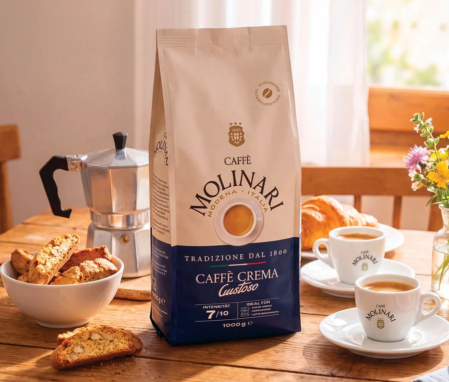 Eine Packung Caffè Molinari Caffè Crema Gustoso Kaffeebohnen steht auf einem Holztisch neben einer silbernen Mokkakanne, einer Schale mit Cantuccini, zwei Tassen Kaffee, Croissants und einem Blumenstrauß.