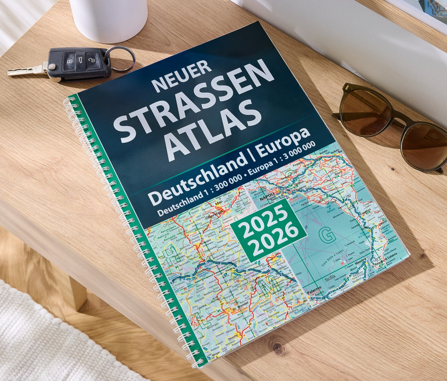 Auf einem Holztisch liegen ein Buch 'Neuer Straßen-Atlas Deutschland/Europa 2025/2026', ein Autoschlüssel und eine Sonnenbrille.