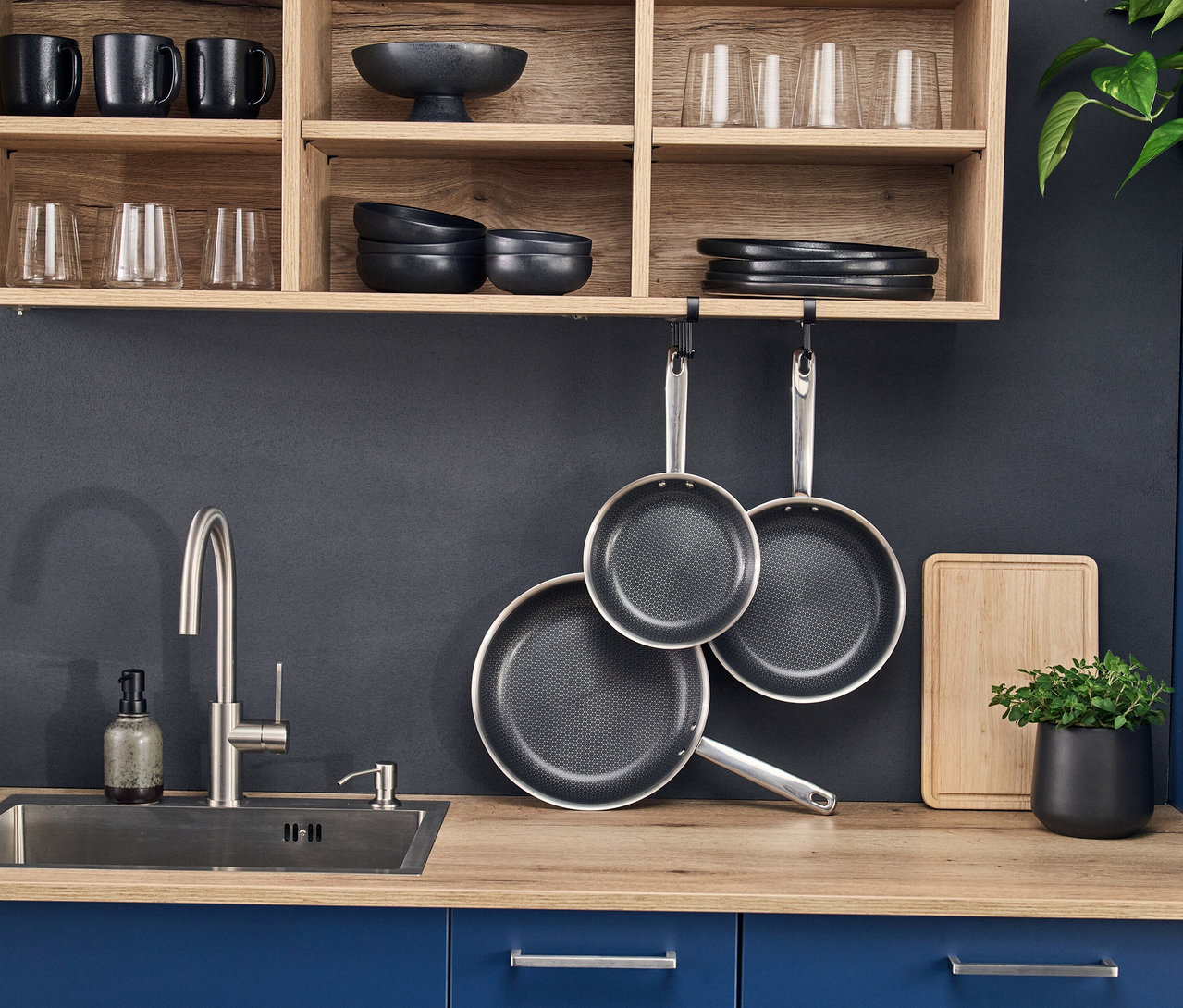 Drei Pfannen des Henckels by ZWILLING »Valley« Bratpfannensets, zwei hängend und eine auf der Arbeitsplatte, in einer modernen Küche mit Spüle, Holzregalen und Küchenutensilien.