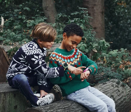 Zwei Jungen sitzen auf Baumstämmen im Wald; der eine Junge zeigt auf die Xplora X6 Play Kids Watch mit grünem Armband am Handgelenk des anderen Jungen.