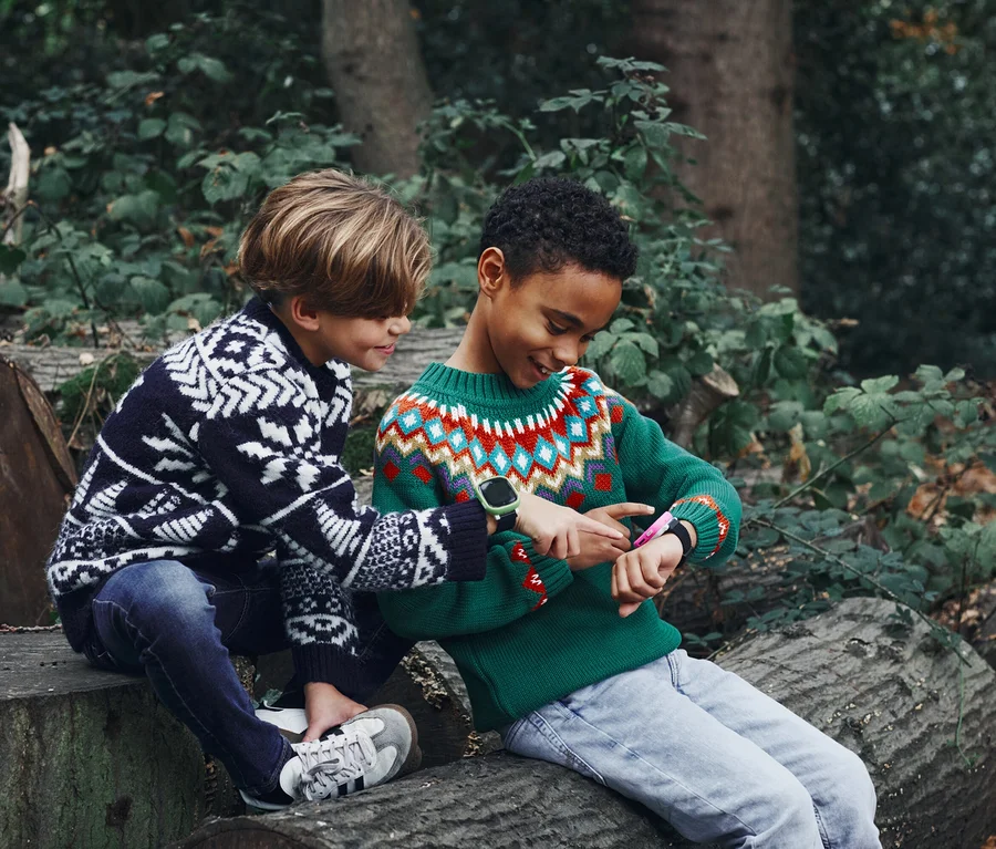 Zwei Jungen sitzen auf Baumstämmen im Wald; der eine Junge zeigt auf die Xplora X6 Play Kids Watch mit grünem Armband am Handgelenk des anderen Jungen.
