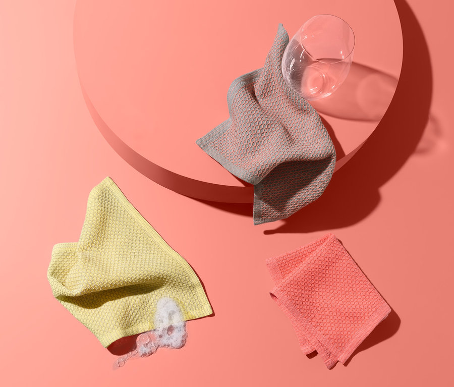 Drei Spültücher liegen auf lachsfarbenem Hintergrund: ein gelbes mit Spülmittelresten, ein rosafarbenes und ein graues auf einem Podest neben einem Glas.