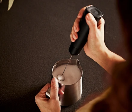 Frau schäumt Milch in einem Edelstahlbehälter mit einem schwarzen Batterie-Milchaufschäumer auf.