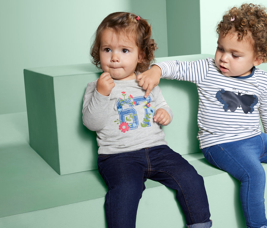 Zwei kleine Kinder sitzen nebeneinander. Eines trägt ein graues Langarmshirt und Jeans, das andere ein gestreiftes Langarmshirt und Jeans.