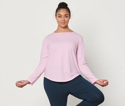 Frau steht in der Baum-Pose auf einer Yogamatte. Sie trägt ein hellrosa Langarm-Sportshirt und eine Yogahose.