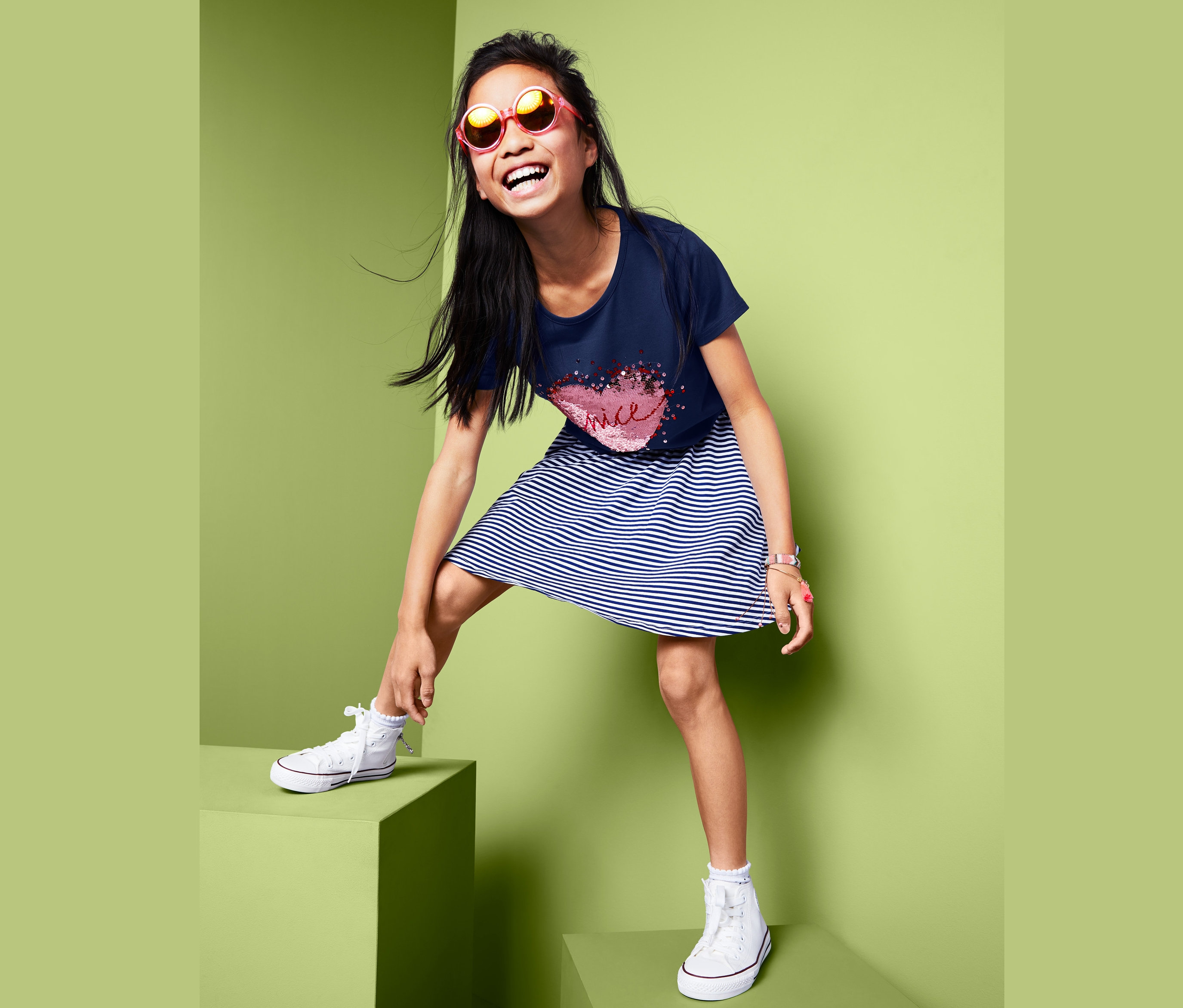 Ein lächelndes Mädchen in einem T-Shirt mit Wendepailletten, einem gestreiften Kleid und Turnschuhen steht auf grünen Würfeln.