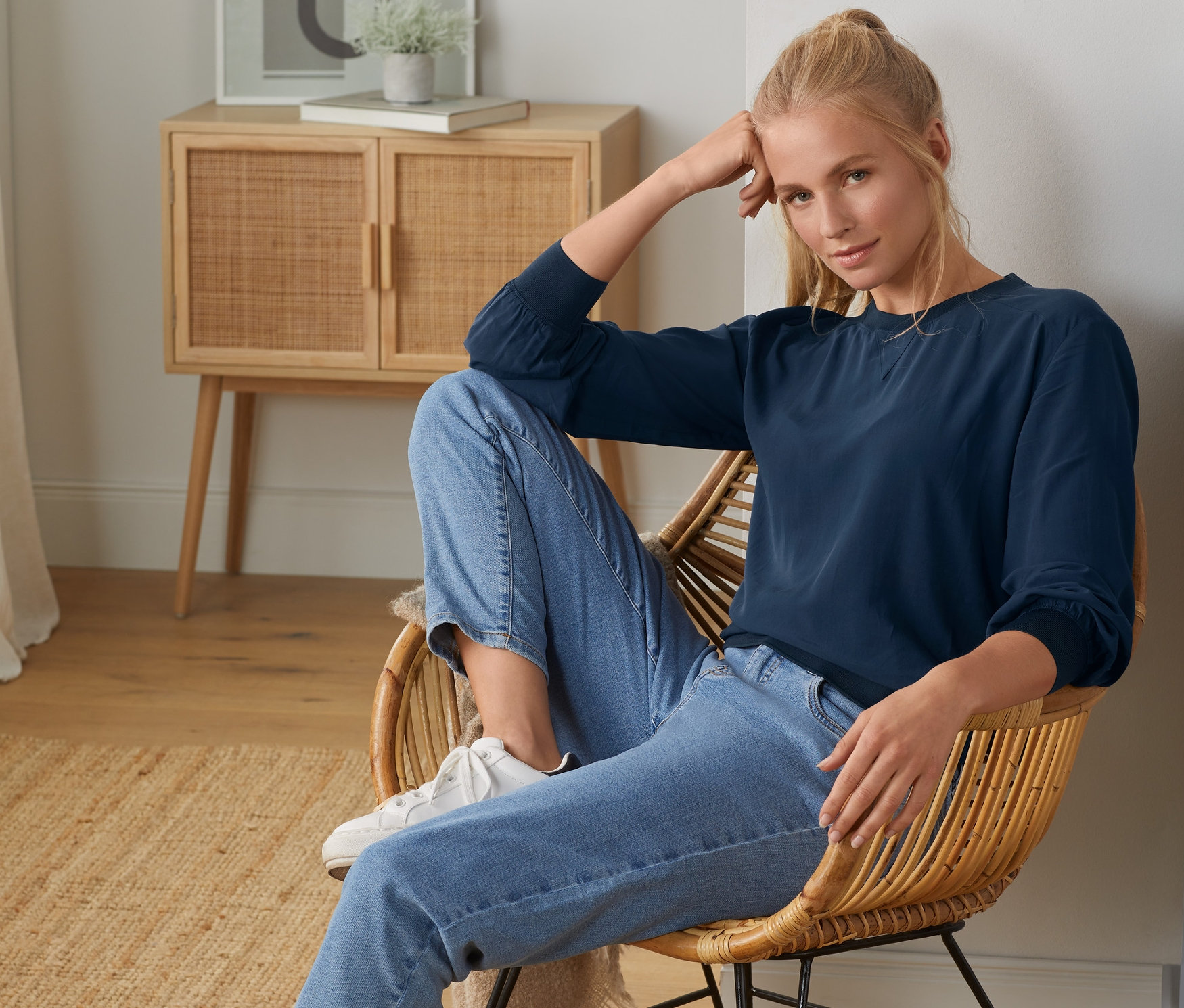 Blonde Frau sitzt im Sessel und trägt eine Bluse mit Rippbündchen.