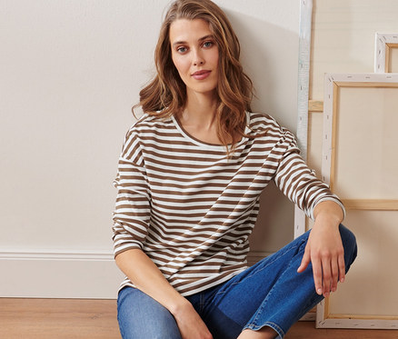 Frau sitzt auf dem Boden neben Leinwänden und trägt ein gestreiftes Langarmshirt, beige.