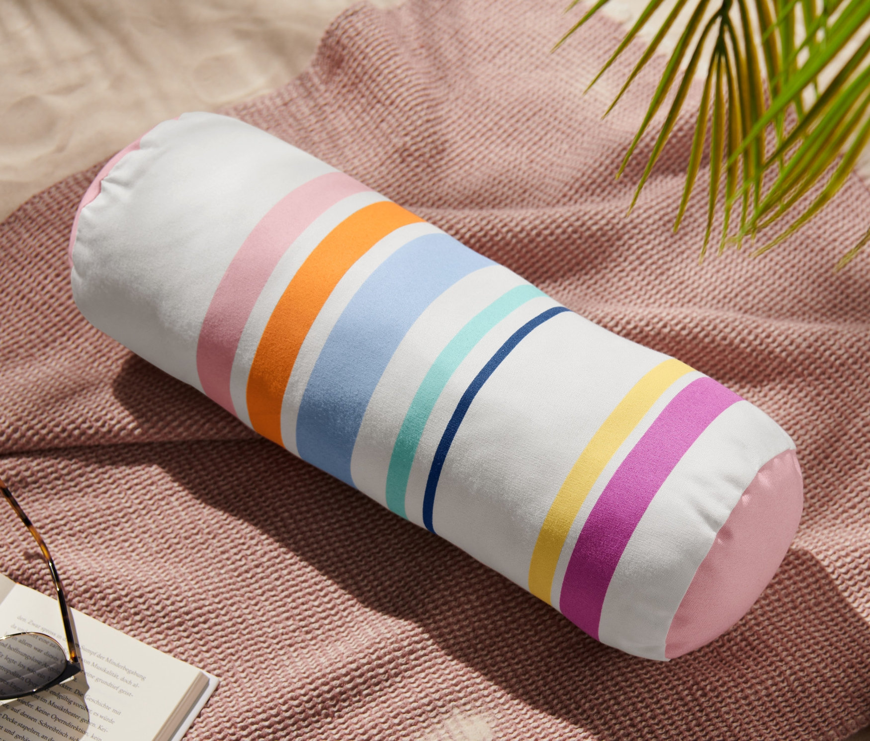 Eine Outdoor-Kissenrolle liegt auf einer rosa Decke neben einem Buch und einer Sonnenbrille.