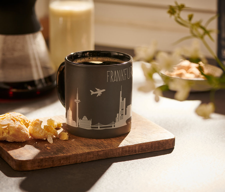 Ein schwarzer Kaffeebecher Frankfurt mit dem Aufdruck "Frankfurt" und einem Flugzeug und Gebäuden steht auf einem Holzbrett neben Keksen.