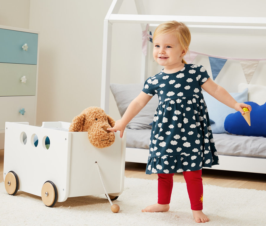 Mädchen in 2 Jerseykleider aus Bio-Baumwolle mit Wolkenmuster steht neben einem weißen Spielzeugwagen mit Plüschhund.