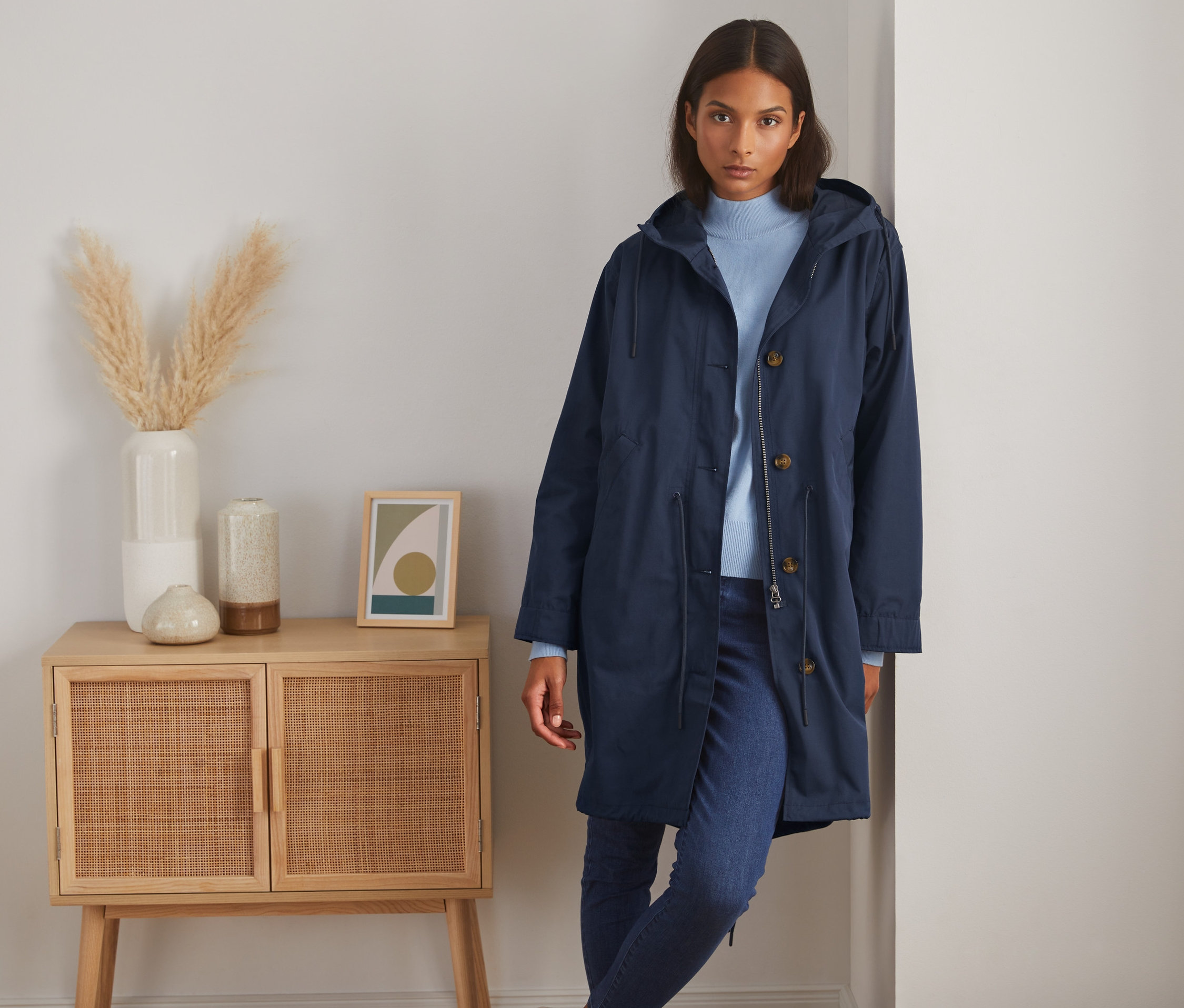 Eine Frau posiert in einem dunkelblauen Parka mit Kapuze neben einem Schrank.