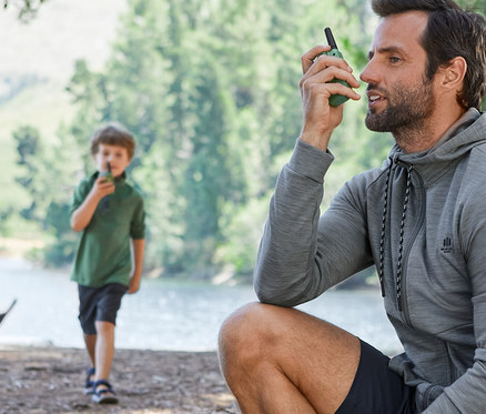 Ein Mann sitzt auf einem Baumstamm und benutzt ein Walkie-Talkie. Im Hintergrund ist ein Junge mit einem Walkie-Talkie zu sehen. Beide tragen Funktionskleidung.