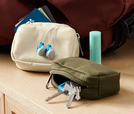 Zwei Mini-Reise-Ordnungstaschen, Kopfhörer und ein Lippenbalsam liegen auf einer Kommode neben einem Rucksack und einer Vase.