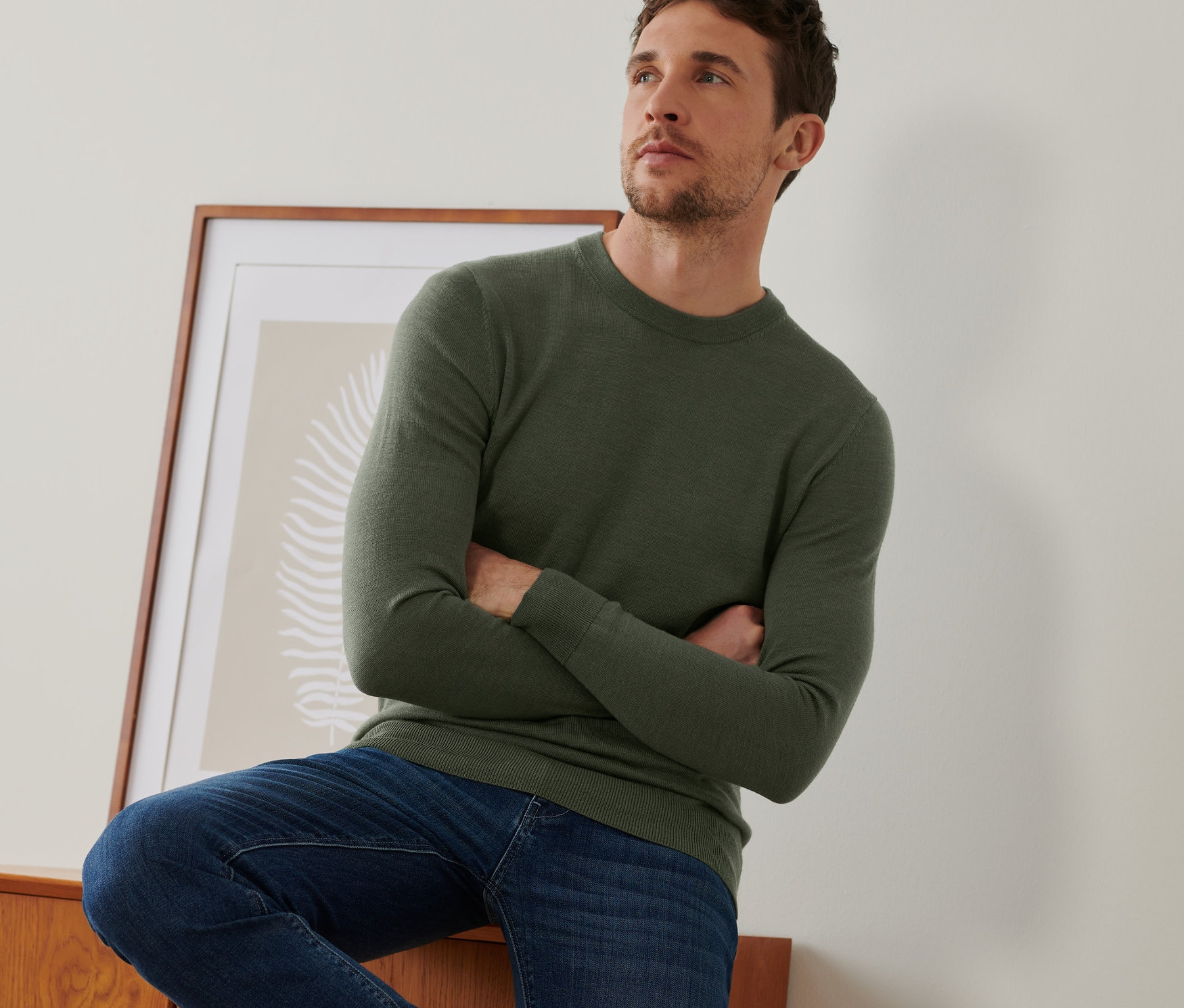 Mann posiert an einer Kommode und trägt einen khakifarbenen Merinopullover.