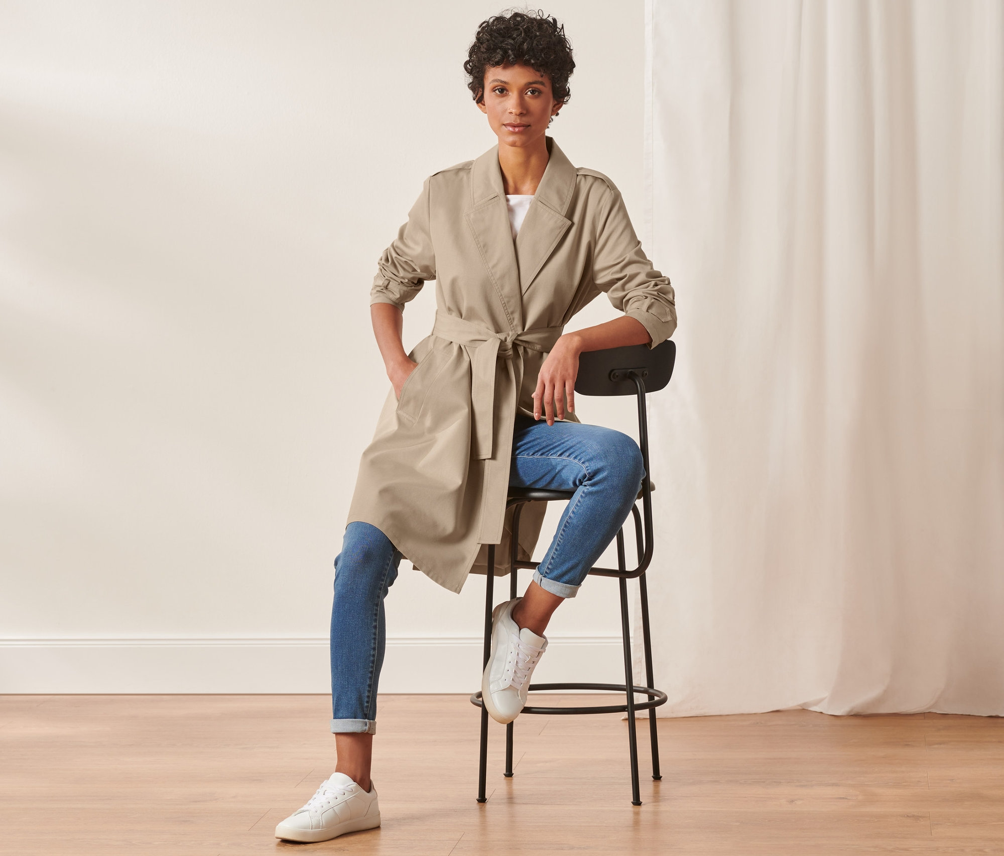 Frau sitzt auf einem Stuhl und trägt einen Trenchcoat, Jeans und Turnschuhe.