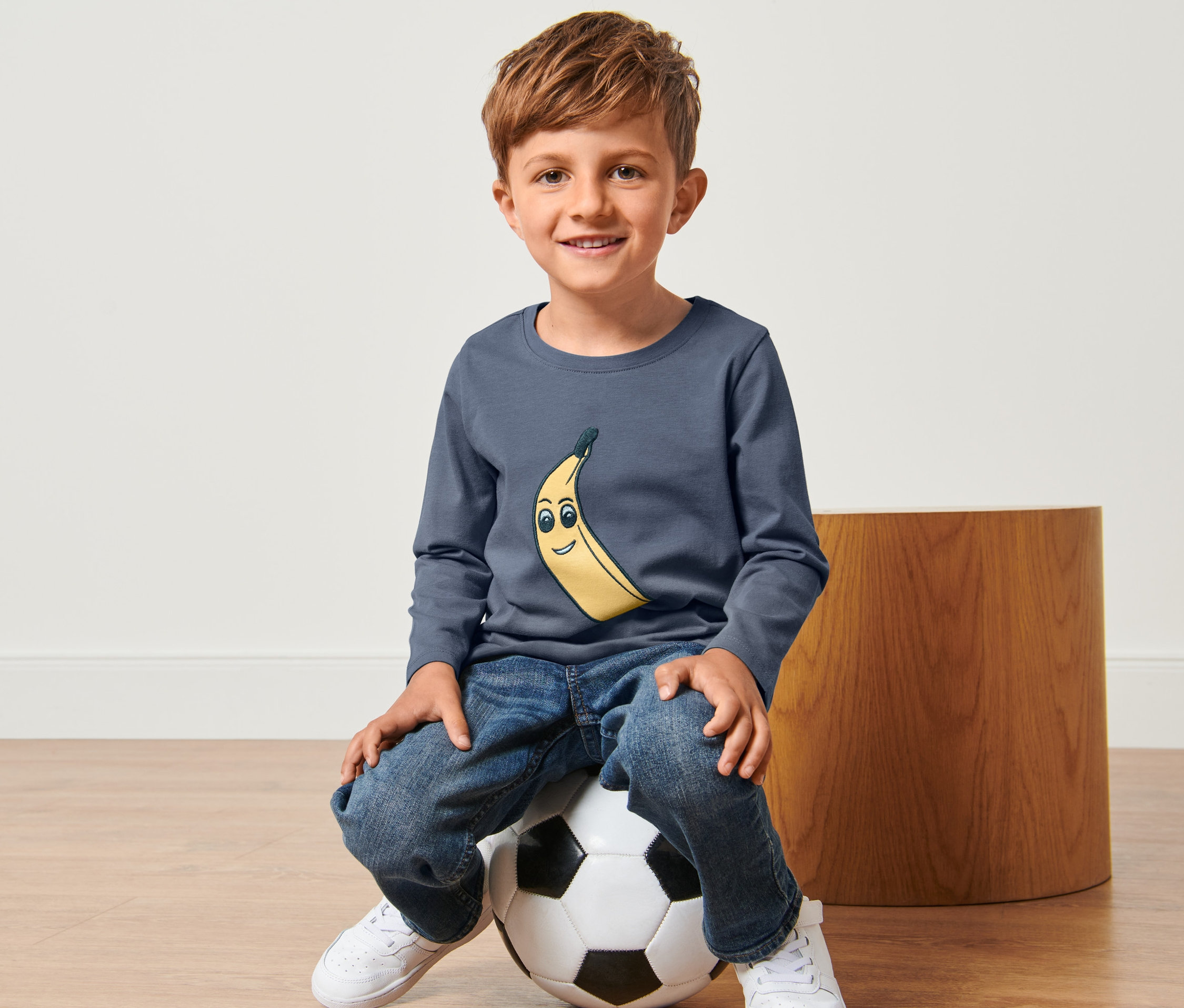 Ein Junge sitzt auf einem Fußball mit einem blauen, langärmeligen Shirt mit Bananenaufdruck, Jeans und weißen Schuhen. Im Hintergrund ist ein Hocker zu sehen. Das Kind trägt ein "2 Kleinkinder-Shirts".
