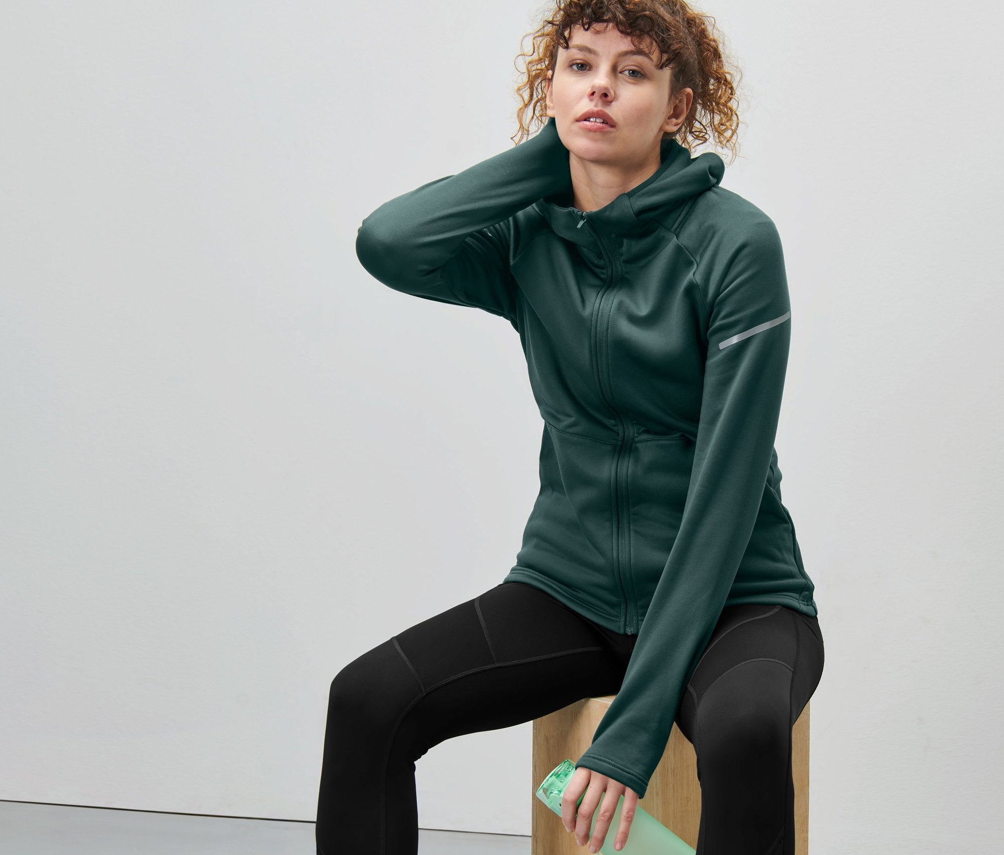 Frau sitzt auf einem Holzblock und trägt eine dunkelgrüne Thermojacke.