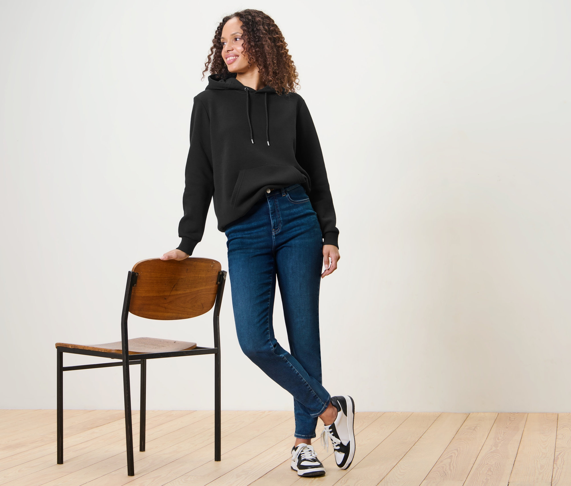 Eine Frau in einem schwarzen Hoodie und blauen Skinny Jeans – Fit »Hanna« lehnt an einem Stuhl.