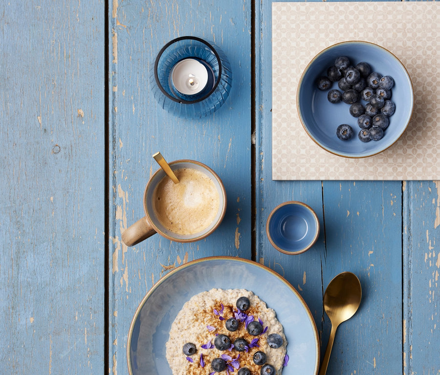 Haferbrei mit Blaubeeren und Blüten, Kaffee, eine Schale Blaubeeren und ein blauer 2 Bitz Teelichthalter »Kusintha« auf einem blauen Holztisch.