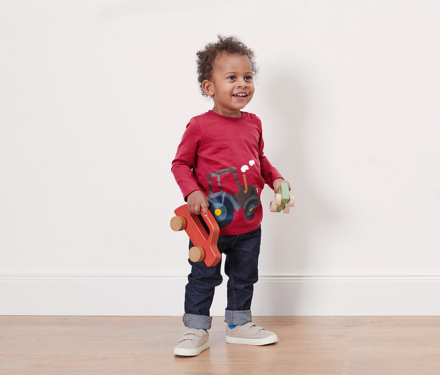 Ein Kleinkind in einem roten Langarmshirt mit Traktor hält zwei Spielzeugautos aus Holz.