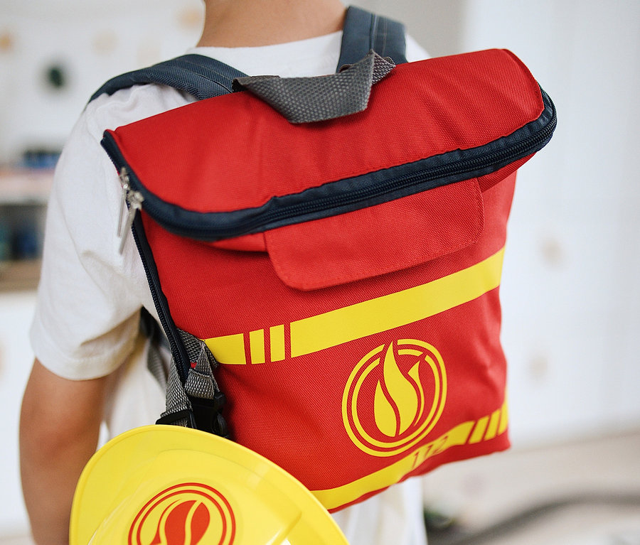 Ein Junge trägt einen roten small foot Feuerwehrrucksack und einen gelben Helm.