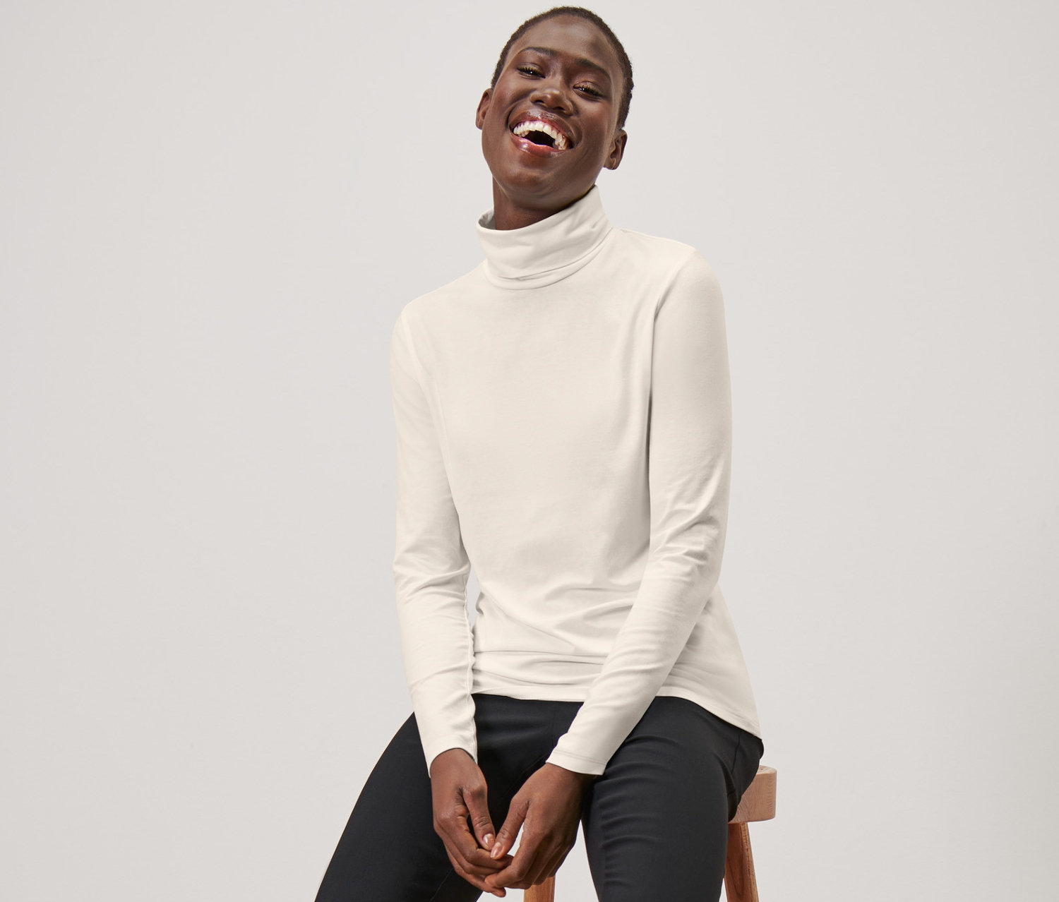 Eine Frau sitzt auf einem Hocker und trägt ein cremeweißes Rollkragenshirt, eine schwarze Hose und schwarze Slipper.