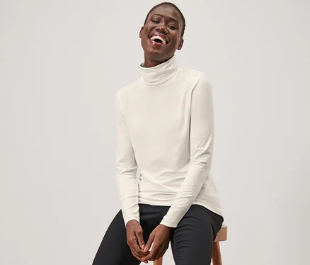 Eine Frau sitzt auf einem Hocker und trägt ein cremeweißes Rollkragenshirt, eine schwarze Hose und schwarze Slipper.